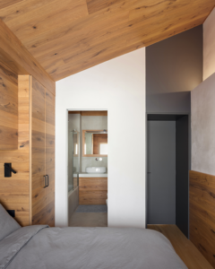 Vista dall'interno della parete di accesso alla camera matrimoniale, da cui si intravede anche il bagno en-suite. Mix di finiture tra laccatura opaca di colore grigio e pareti e soffitto rivestiti in quercia.