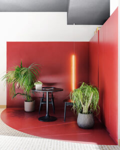 Angolo lounge dal design contemporaneo caratterizzato da pareti e pavimento rosso intenso, con tavolino rotondo e sedie nere minimal. Due piante verdi in vaso aggiungono un contrasto naturale, mentre una luce verticale a parete crea un’atmosfera calda e accogliente.