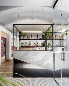 Bancone bar dal design contemporaneo con struttura in metallo nero e rivestimento in marmo, arricchito da mensole con bottiglie a vista e piante decorative. Lo spazio è luminoso e moderno, con dettagli in vetro e linee pulite che valorizzano l’ambiente.