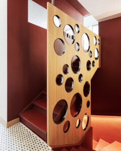 Scala interna dal design contemporaneo con pareti rosso intenso e una parete divisoria in legno chiaro traforata da aperture circolari di diverse dimensioni, che lasciano intravedere i gradini e gli spazi retrostanti; illuminazione calda integrata lungo i bordi accentua le linee architettoniche.