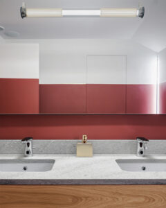 Bagno moderno con doppio lavabo incassato in piano in pietra chiara, rubinetti cromati e dispenser centrale; parete bicolore bianco e rosso con specchio integrato e illuminazione lineare a soffitto dal design contemporaneo.