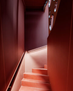 Scala interna moderna con pareti rosso intenso e gradini coordinati, illuminata da una sottile luce lineare lungo la parete; corrimano scuro a contrasto e porzione di pannello divisorio in legno con aperture circolari visibile sul lato superiore.