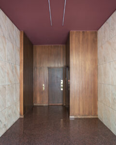 Un corridoio dal design moderno ed elegante, caratterizzato da una combinazione di pareti rivestite in pannelli di legno, piastrelle effetto marmo e un pavimento lucido dalle tonalità rossastre. Il soffitto è dipinto in un color bordeaux tenue ed è dotato di illuminazione lineare incassata. Una porta è posizionata centralmente alla fine del corridoio.