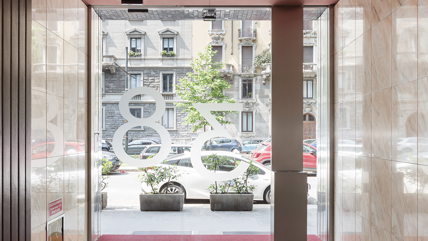 Vista dall'interno dell'androne verso l'esterno. Parete totalmente vetrata con vetrofania con il numero civico e cassetta della posta in acciaio satinato