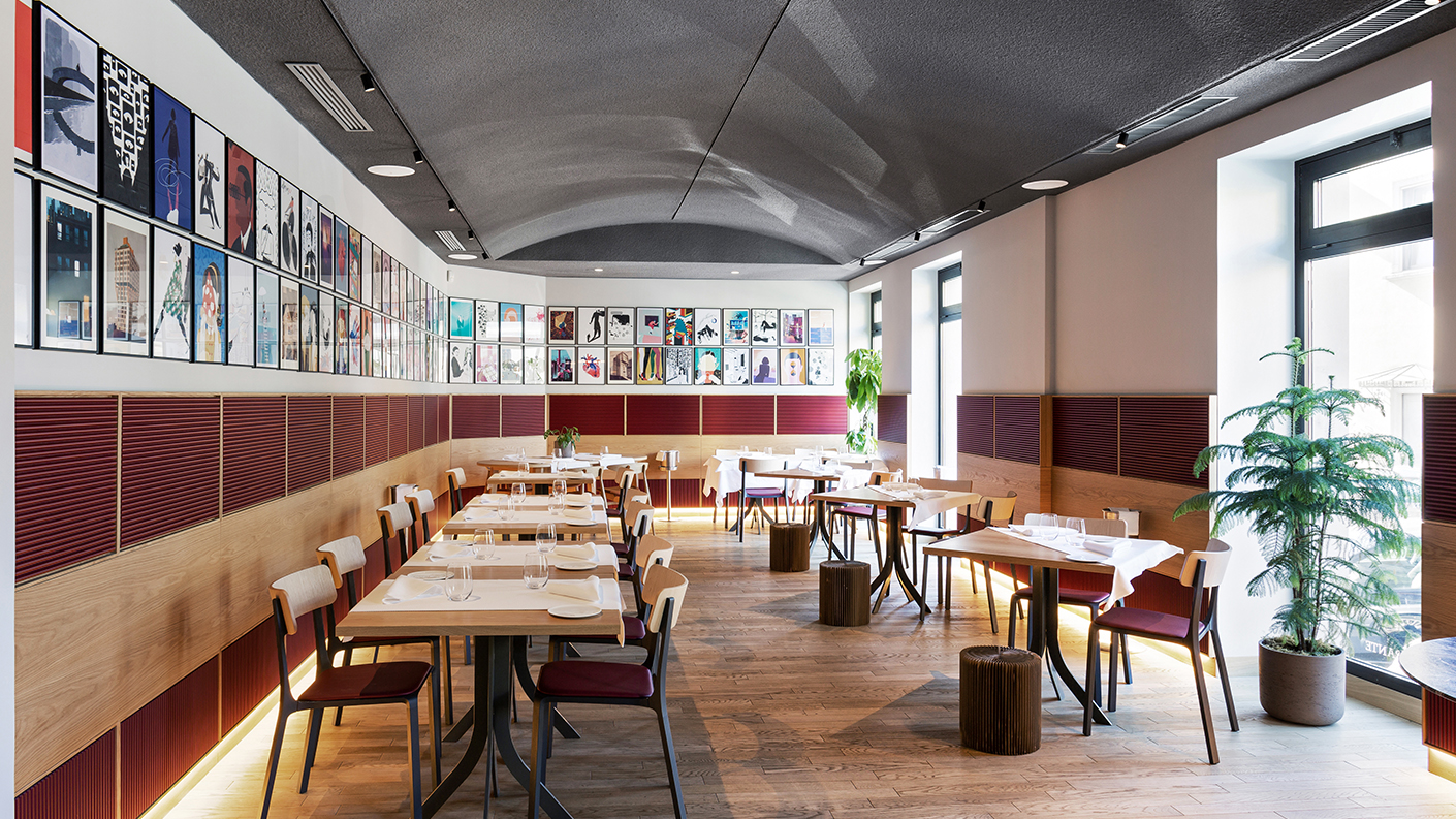 Interno di un ristorante moderno con una fila di tavoli e sedie disposti ordinatamente. Le pareti sono decorate con opere d’arte colorate incorniciate, allineate in un’unica linea lungo tutta la lunghezza della sala. L’arredo combina elementi in legno e accenti rossi, mentre piante in vaso aggiungono un tocco di verde accanto alle finestre. Il soffitto, caratterizzato da una forma ad arco, crea un’atmosfera aperta e accogliente. La luce naturale entra dalle ampie finestre, valorizzando il design pulito e contemporaneo.